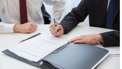 A Person in Black Suit Holding a Pen Near the Documents on the Table